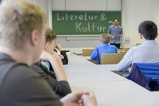 Jungen sitzen am Tisch und ein Mann steht an einer Tafel und erzählt etwas über Literatur und Kultur