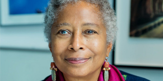 Portrait von Alice Walker: eine Schwarze Frau mit kurzen, grauen Haaren. Sie trägt eine dunkle Bluse mit lilanem Halstuch und hält eine Orangene Rose in der Hand.
