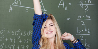 Ein Mädchen steht vor einer Tafel mit mathematischen Formeln. Sie reckt einen Arm in die Höhe. Sie freut sich.
